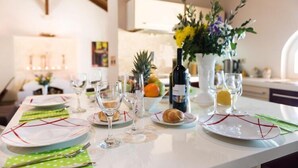 Una mesa de comedor preparada con platos, cubiertos, vasos y una botella de vino, con un florero de flores y frutas al fondo.