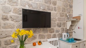 A stone wall with a flat-screen TV mounted above a white table with a vase of yellow flowers and two oranges.