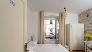 A hotel room with a bed, a wardrobe, a window with curtains, a small table with a vase of flowers, and a view of the outside balcony.