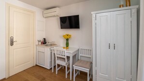 Una habitación pequeña y limpia, con una puerta blanca, una mesa blanca y dos sillas, un televisor de pantalla plana montado en la pared, y un armario blanco con dos puertas.