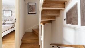 Una escalera con escalones de madera y una pared blanca. En la habitación contigua, hay una cama con ropa de cama blanca.