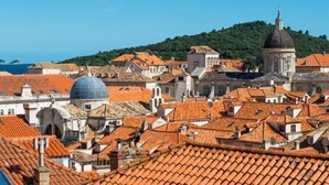 A coastal town with terracotta-roofed buildings, a prominent dome, and a hill in the background.