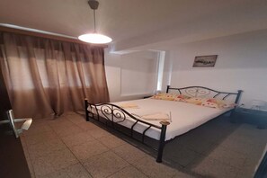 A single bed with a metal frame, a bedside table, a hanging light fixture, a window with curtains, and a wall-mounted picture.