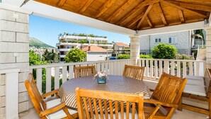 A covered outdoor seating area with a round table and wooden chairs, overlooking a residential area with buildings and greenery. - Apartment Vulicevic A1 (Dubrovnik)