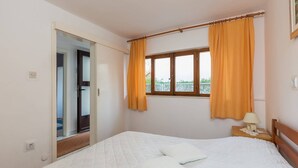 A bedroom with a bed, two pillows, a nightstand with a lamp, a window with curtains, and a door leading to another room.