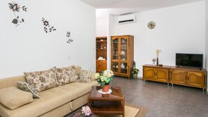 Una sala de estar con un sofá color beige, un aparador de madera, un televisor y una mesita con un jarrón de flores.