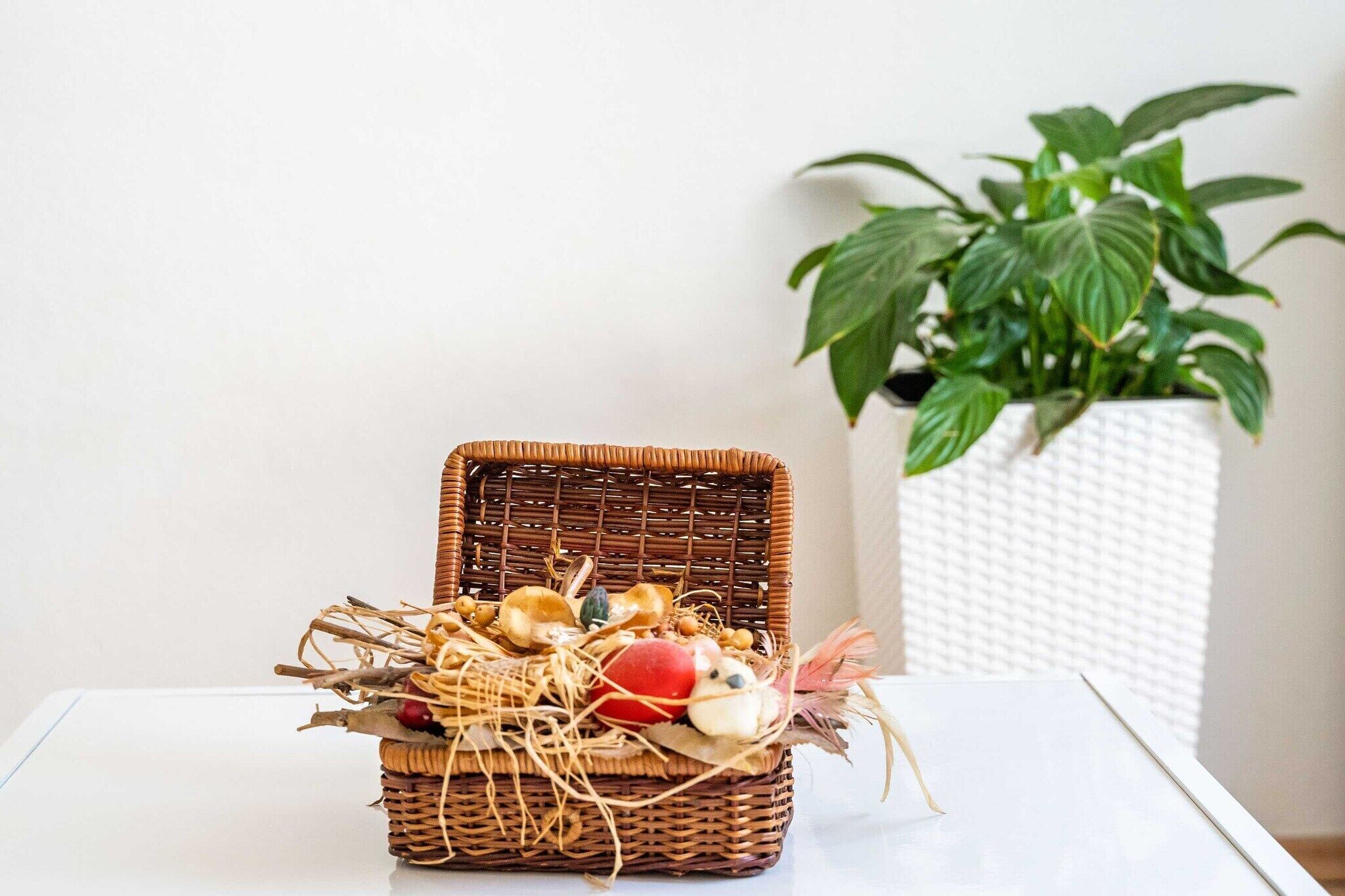 Un panier en osier rempli d’objets décoratifs est posé sur une table blanche, à côté d’un pot blanc texturé dans lequel se trouve une plante verte.