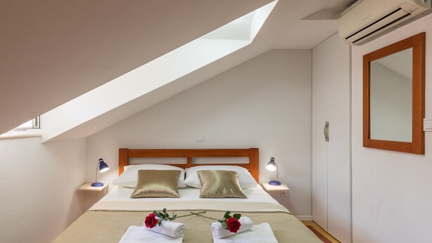 A neatly arranged bedroom with a sloped ceiling, a bed with two pillows, bedside lamps, a wardrobe, and a mirror. - Apartments Ivana Old Town - Standard Studio - APT3 (Dubrovnik)