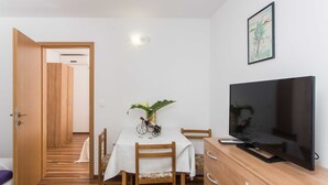 A room with a wooden floor, a dining table with chairs, a TV on a wooden cabinet, and a door leading to another room.