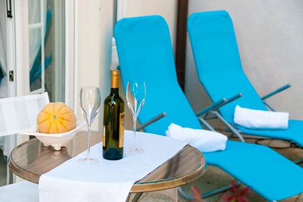 A table with a bottle of wine, two glasses, and a bowl with a fruit. Blue lounge chairs are in the background.