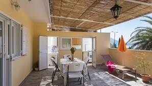 A sunlit patio with a table and chairs, a small table with a vase, a chair with a pink cover, and a potted plant. - Apartments Djurkovic - Two-Bedroom Apartment with Shared Terrace and Sea View (Mlini)