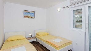 A small, clean room with two single beds, a bedside table with a lamp, a painting on the wall, and a window with blinds. - Apartments Djurkovic - Two-Bedroom Apartment with Shared Terrace and Sea View (Mlini)