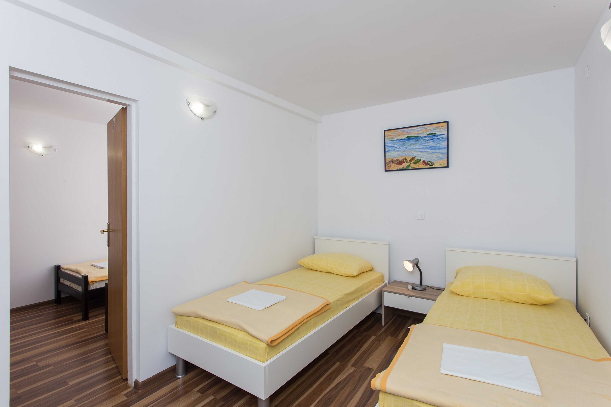 Une chambre avec deux lits simples, un plancher en bois, un tableau au mur et une porte donnant sur une autre pièce.
