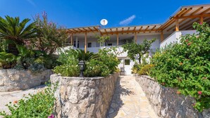 A stone pathway leading to a white building with a wooden pergola, surrounded by lush greenery and flowering plants. - Apartments Djurkovic - Two-Bedroom Apartment with Shared Terrace and Sea View (Mlini)