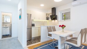 Una cocina moderna con armarios blancos, un refrigerador de acero inoxidable y un microondas. Hay un comedor con una mesa y sillas blancas, y un florero con flores rojas sobre la mesa.