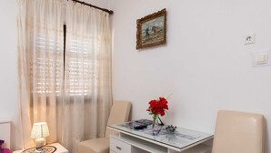 Ein Zimmer mit Schreibtisch mit Glasplatte, einem Stuhl, einer Lampe, einer Vase mit roten Blumen, einem gerahmten Bild an der Wand und einem Vorhang.