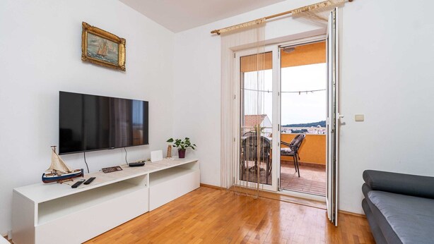 A living room with a flat-screen TV, a white entertainment unit, a balcony with a table and chairs, and a grey sofa.