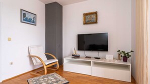 A modern living room with a flat-screen TV, a white sofa, a wooden chair, a small table, and two framed pictures on the wall.