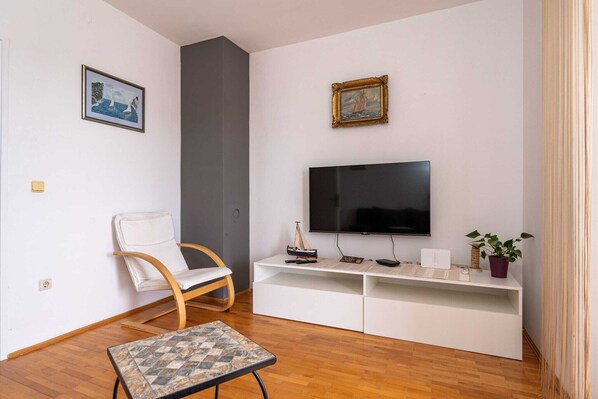 En moderne stue med et fladskærms-tv, en hvid sofa, en træstol, et lille bord og to billeder i rammer på væggen.