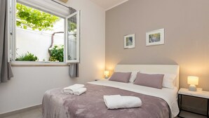 A neatly made bed with a white and purple bedspread, two bedside lamps, and a window with a view of greenery. - Rooms Batina - Double Room-1 (Dubrovnik)