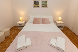 A bedroom with a bed, two bedside tables with lamps, a pair of pillows, a wooden floor, and two framed pictures on the wall.