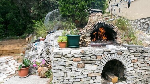 Un four extérieur en pierre, dans lequel brûle un feu, est entouré de plantes en pot et d’un mur de pierre.