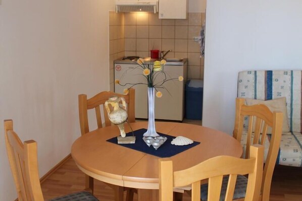 A small dining area with a round wooden table, four chairs, a vase with flowers, and a decorative shell. A kitchenette with a stove and cabinets is visible in the background.