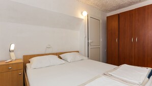 Una cama bien hecha con sábanas blancas, una mesita de noche de madera con una lámpara, y un armario con espejo.