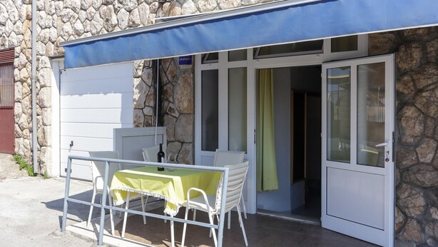 Un pequeño espacio al aire libre con una mesa, sillas, un muro de piedra y un toldo azul.