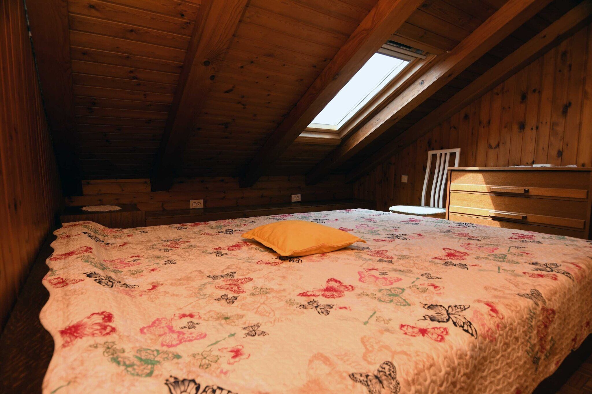 Une chambre avec un plafond en bois, une lucarne, un lit recouvert d’une couvre-lit à motifs floraux et une commode en bois.