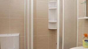 Un baño con azulejos color beige, inodoro blanco, cabina de ducha de cristal y lavamanos con una botella de jabón de manos Generica.