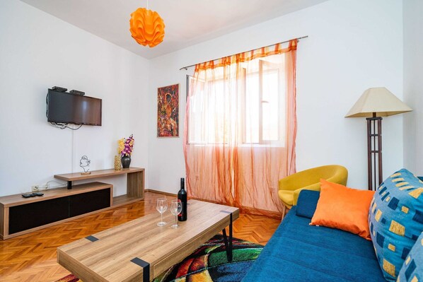 A modern living room with a wooden coffee table, a blue sofa with orange and blue cushions, a yellow armchair, a floor lamp, a TV mounted on the wall, and a large window with sheer curtains.