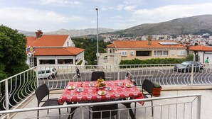 A terrace with a table set for dining, overlooking a residential area with red-roofed houses and a hilly landscape.