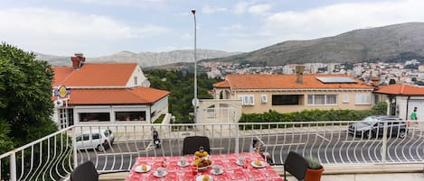 A terrace with a table set for dining, overlooking a residential area with red-roofed houses and a hilly landscape.