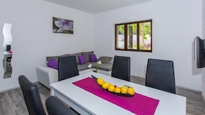A modern living room with a dining area, featuring a sofa, chairs, a table with a fruit centerpiece, and a large window with a view of greenery. - Villa Roza- Two-Bedroom Apartment (Mokosica)