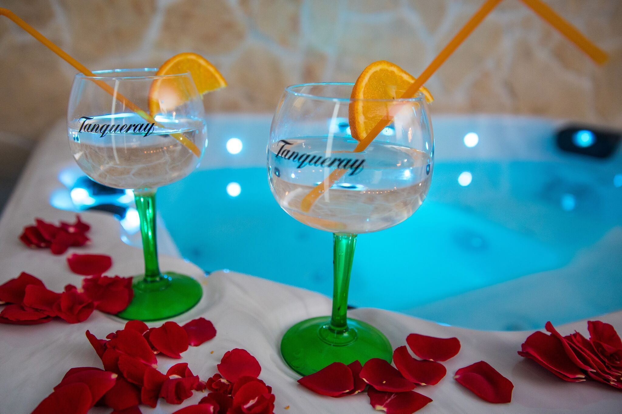 Two glasses of a clear beverage with orange slices and straws, labeled "Tanqueray," on a table with red rose petals.