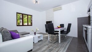A modern kitchen with white cabinets, a dining area with a white table and black chairs, and a gray sofa with a purple pillow. - Villa Roza- Two-Bedroom Apartment (Mokosica)
