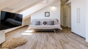 A modern loft bedroom with a sloped ceiling, a flat-screen TV, a sofa with cushions, and a framed picture on the wall.