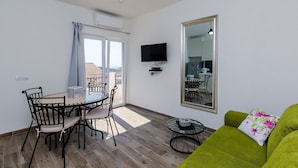 A living room with a green sofa, a dining table with chairs, a TV mounted on the wall, and a large mirror. - Apartments Life - One Bedroom Apartment with Balcony and Sea View (Dubrovnik)