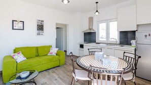 A compact living space with a green sofa, a round dining table with chairs, a kitchen area with white cabinets and a stove, and a small window. - Apartments Life - One Bedroom Apartment with Balcony and Sea View (Dubrovnik)