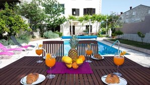 Ein Holztisch mit Frühstücksangeboten und Getränken, ein Schwimmbecken und ein Gebäude im Hintergrund.