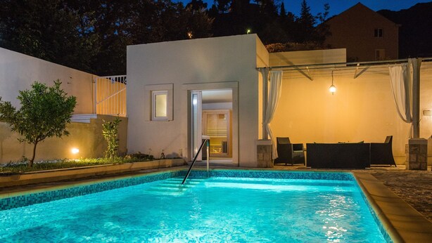 Ein Swimmingpool mit klar blauem Wasser, umgeben von einem weißen Gebäude mit Glastür und einer überdachten Terrasse mit Gartenmöbeln.