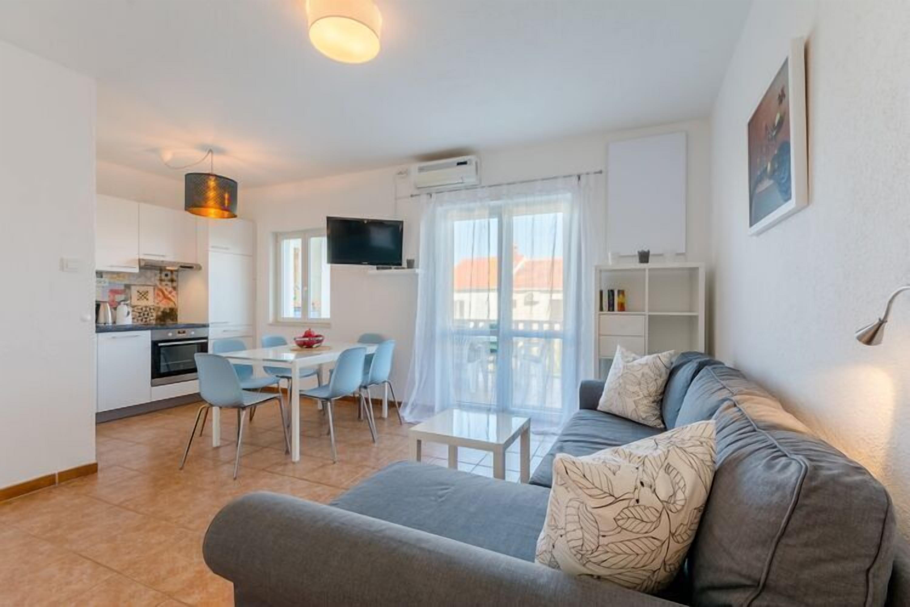 A modern living room with a grey sofa, a dining table with chairs, a kitchen area, and a balcony with a view.