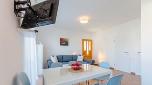 Ein modernes Wohnzimmer mit einem weißen Sofa, blauen Sesseln, einem weißen Tisch mit Obstschale und einem an der Wand befestigten Fernseher.