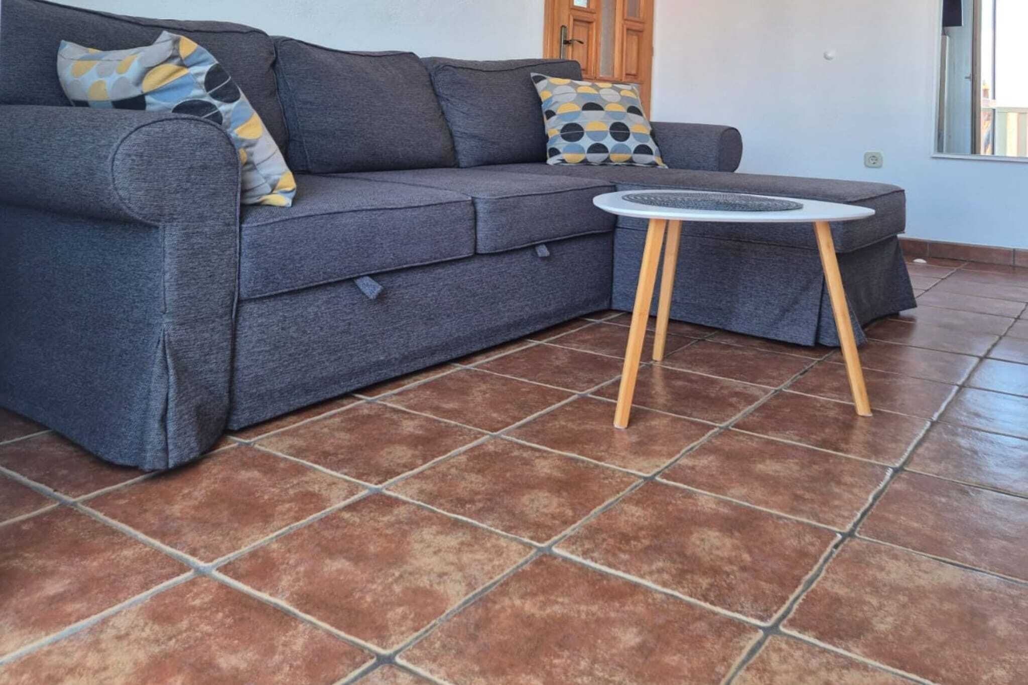 A grey sofa with patterned cushions, a small round table with wooden legs, and a tiled floor.