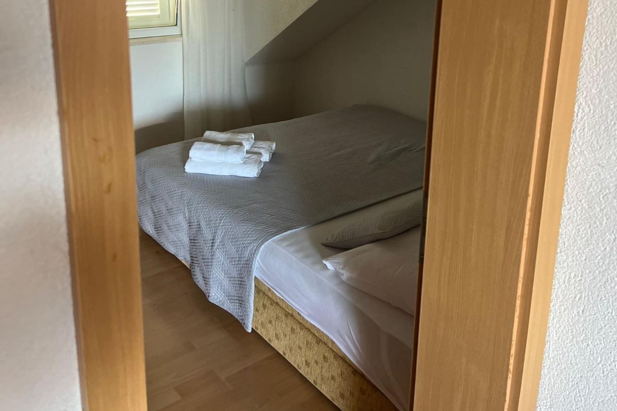 Une petite chambre à coucher, chaleureuse et accueillante, avec un lit simple, une tête de lit en bois et une fenêtre équipée de stores.