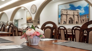 A dining area with a table set for two, featuring a vase with flowers and a painting of a historical building on the wall.