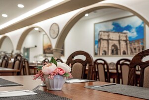 A dining area with a table set for two, featuring a vase with flowers and a painting of a historical building on the wall.