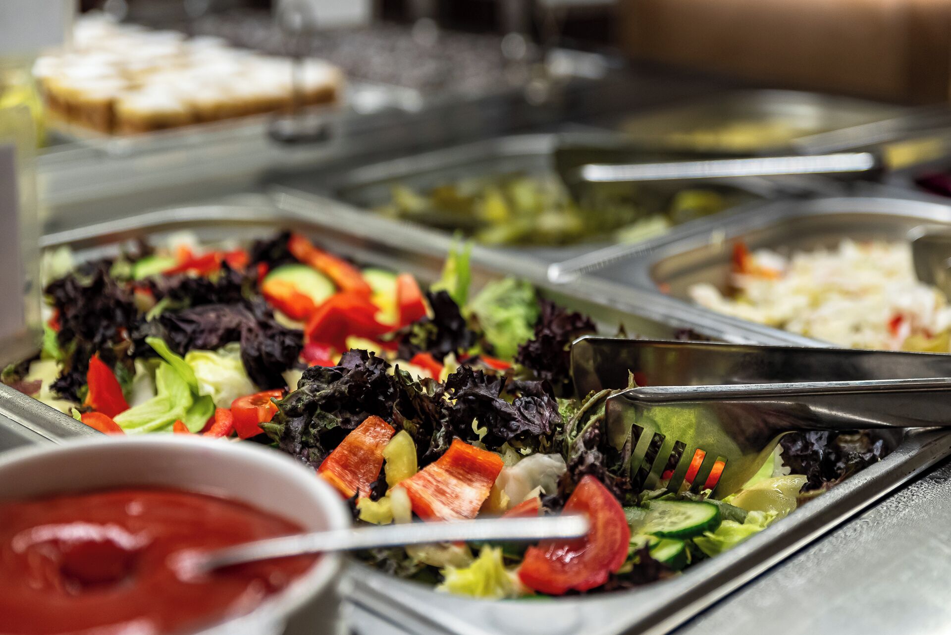 Un bufé con diversas opciones de comida, incluyendo ensaladas, verduras y un bol de salsa roja.