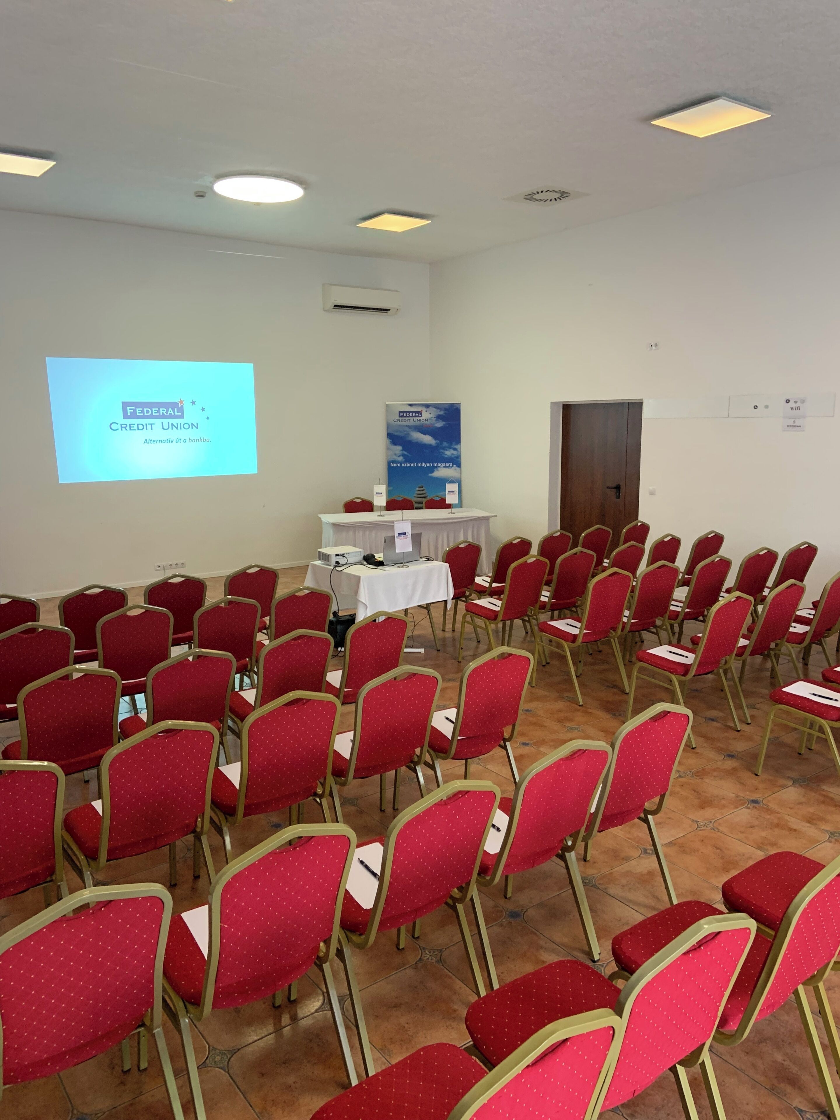A conference room with rows of red chairs, a white table with a projector screen displaying 'Federal Credit Union', and a banner on the wall.